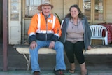 Lester and Val Cain sit on a bench outside an old pub with a sign that reads "Middleton Hotel: Beer, Food, Fuel, Camp"