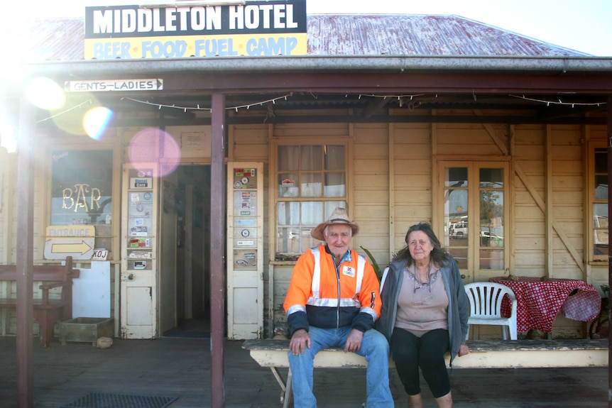 Lester and Val Cain sit on a bench outside an old pub with a sign that reads "Middleton Hotel: Beer, Food, Fuel, Camp"