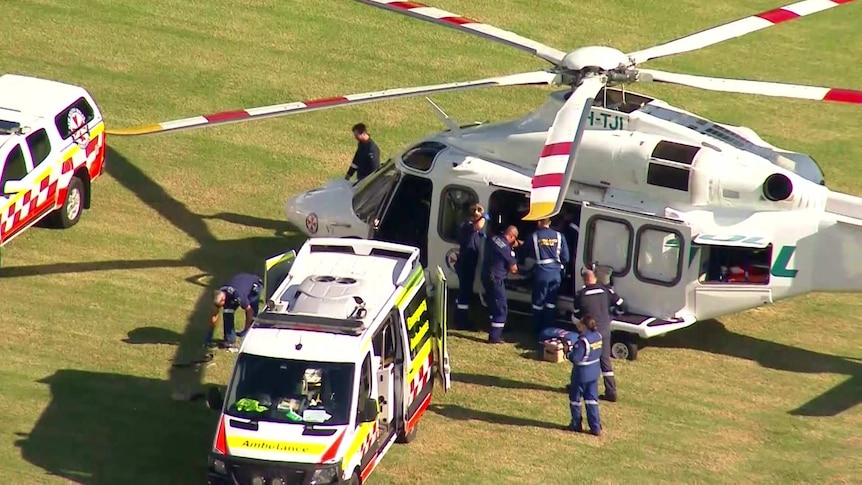 Several people crowd over a stretcher near two large cars and a helicopter in a large field