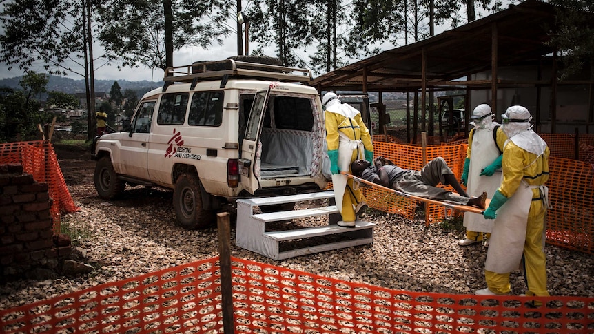 Three people in Ebola scrubs carry a patient on a stretched into an ambulance.