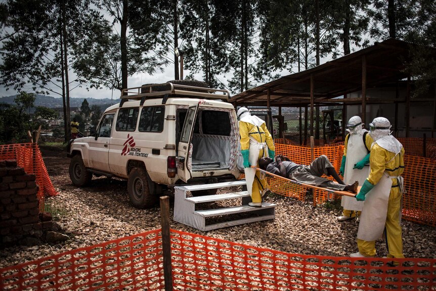 Three people in Ebola scrubs carry a patient on a stretched into an ambulance.