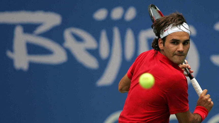 A headband-wearing Olympics men's tennis player focuses on ball as he prepares to hit a backhand.