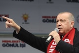Wearing a red scarf and a black jacket, Turkish leader Tayyip Erdogan points while speaking into a microphone.
