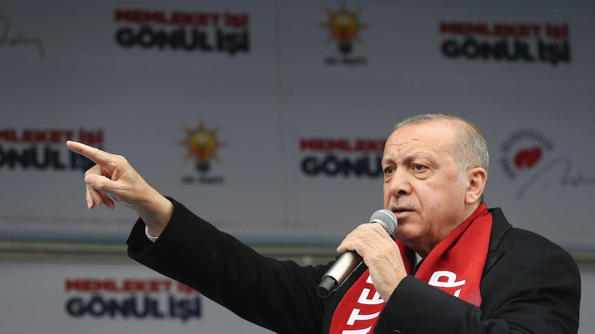 Wearing a red scarf and a black jacket, Turkish leader Tayyip Erdogan points while speaking into a microphone.