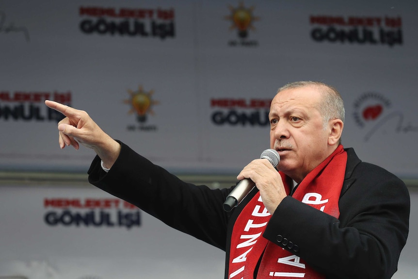 Wearing a red scarf and a black jacket, Turkish leader Tayyip Erdogan points while speaking into a microphone.