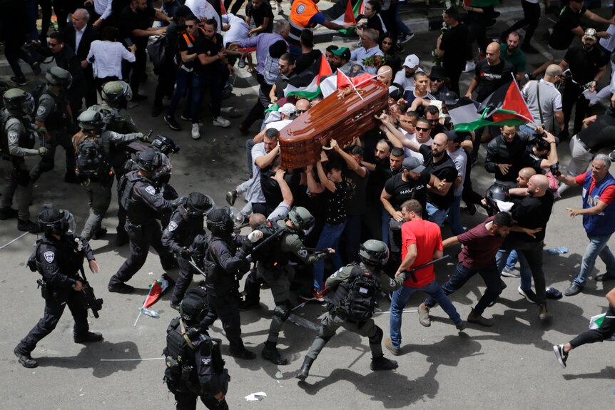 A group of riot police in black riot gear advance on and beat young Palestinian men in civilian clothes carrying a coffin.