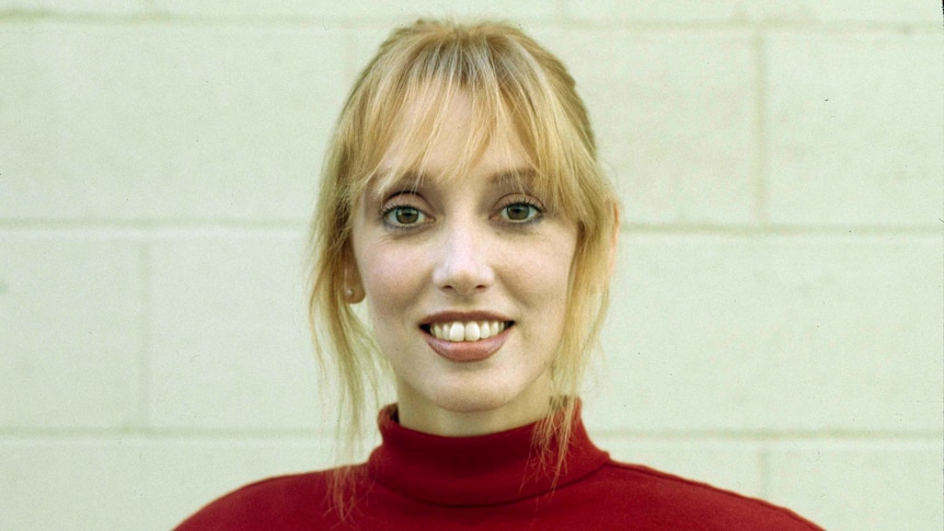 Shelley Duvall with short blonde hair and a fringe smiles at the camera wearing a red shirt