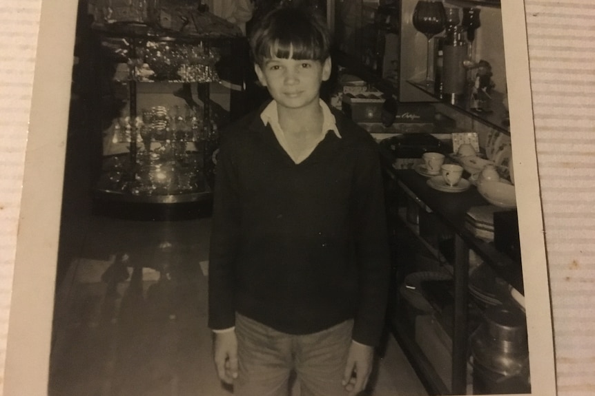 Black and white photo of young box smiling, wearing jumper and shorts, in a room filled with shelves of dinnerware. 
