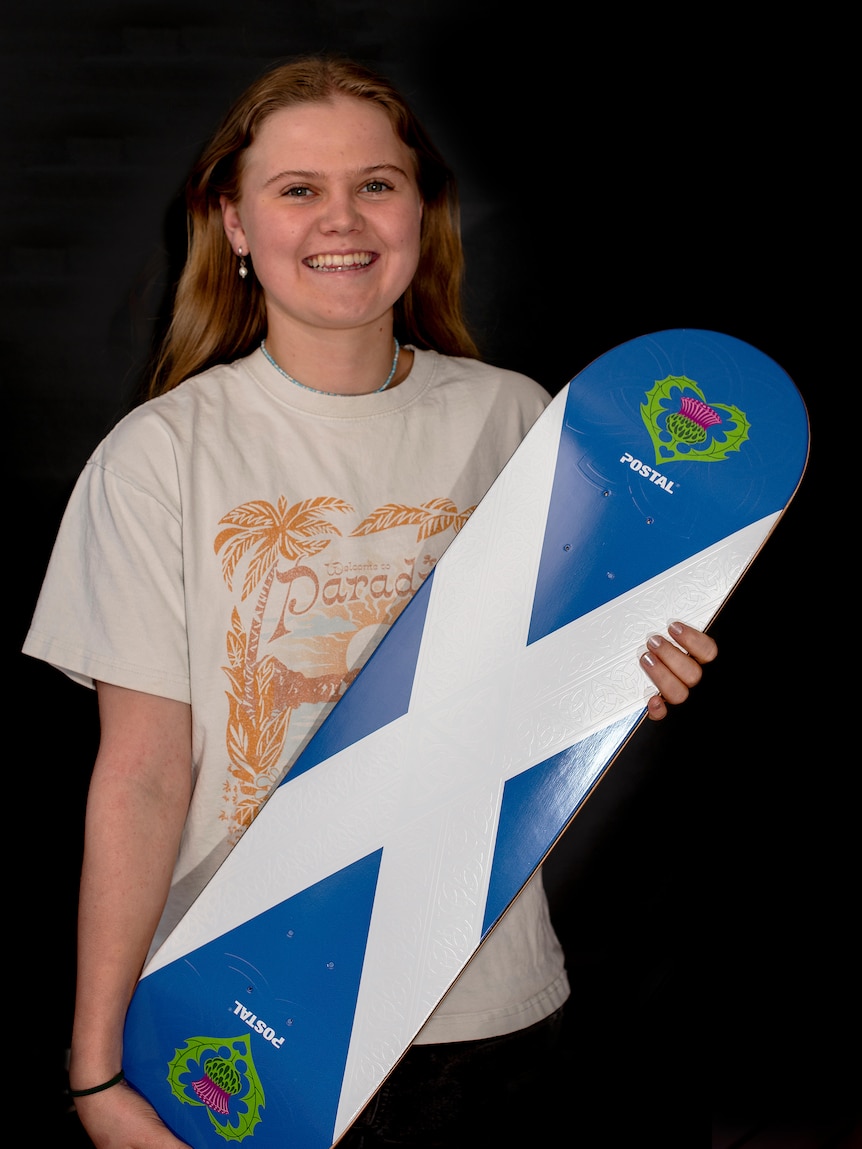 Katie Pike smiling while holding a blue and white skateboard deck. 