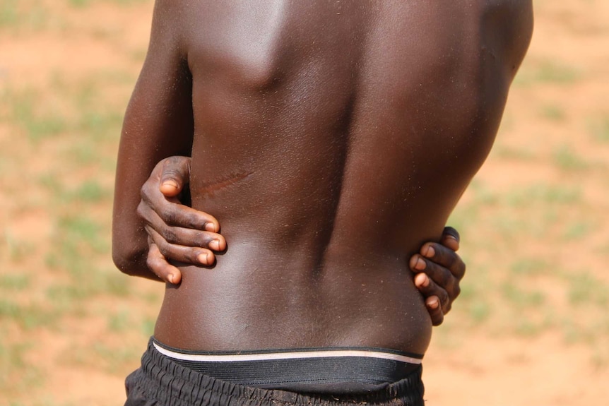 The back of a shirtless Aboriginal teenage boy wearing black pants and with his arms wrapped around his waist.