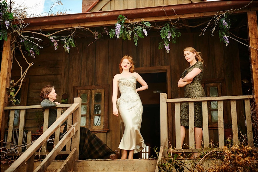 Three women on the verandah of a wooden house, one wearing a formal dress.