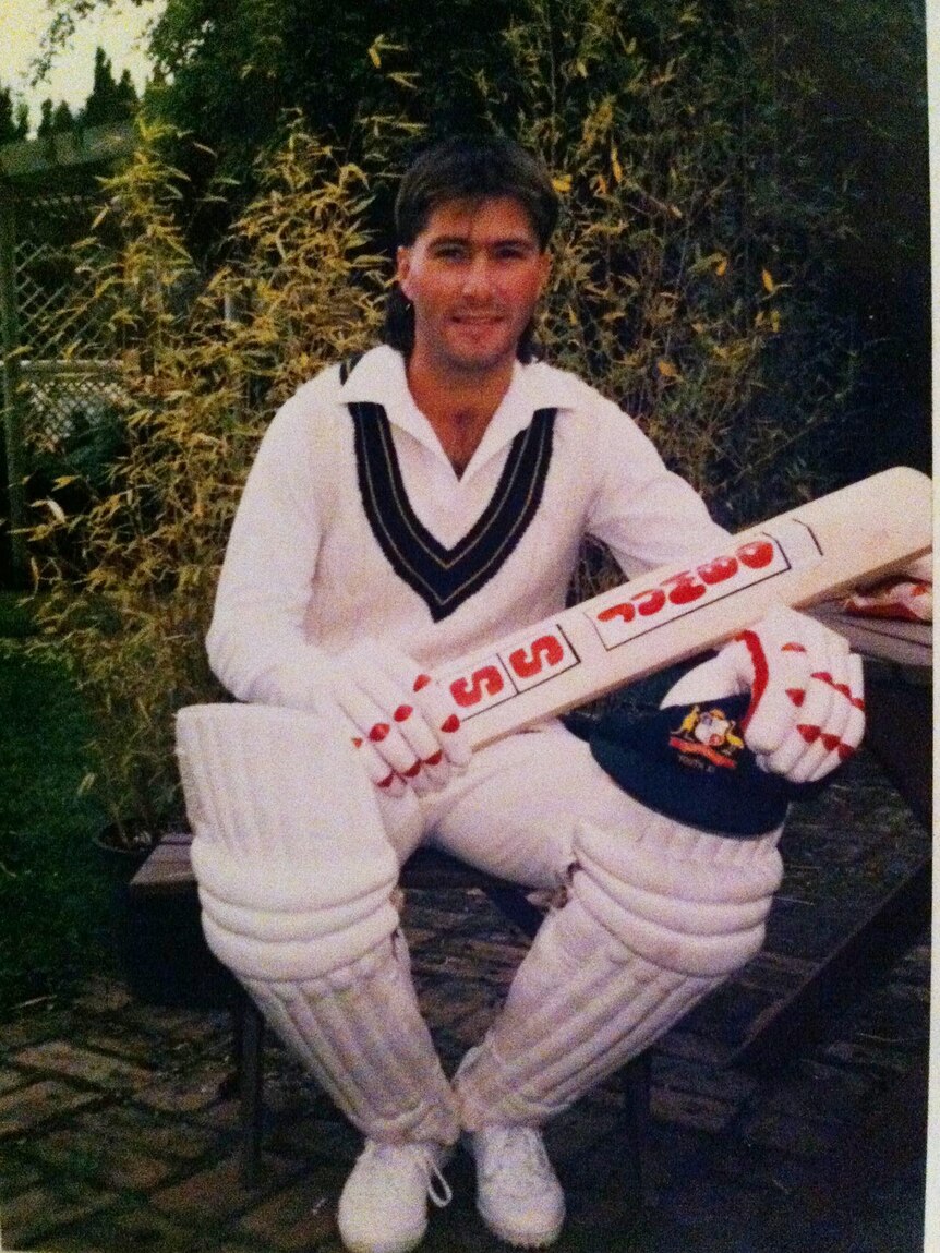 Jamie Mitchell sits in a yard, holding a bat and dressed in full cricket gear with a cap on his knee.