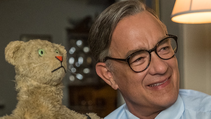An older man in shirt and tie with greying hair and glasses sits on couch and smiles while controlling brown tiger puppet.