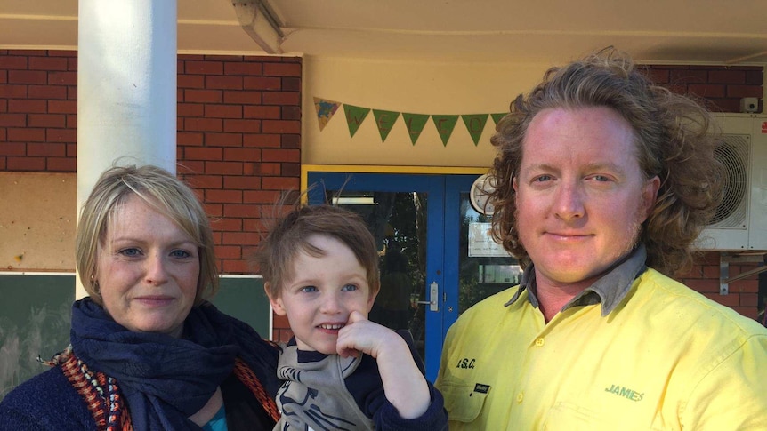 A blonde woman in dark blue jumper and scarf holding young boy standing next to curly haired male in yellow shirt