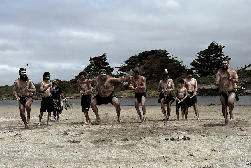 A group of indigenous men from Gunditjmara country perform dreaming ceremony on the beach with trees in the background 