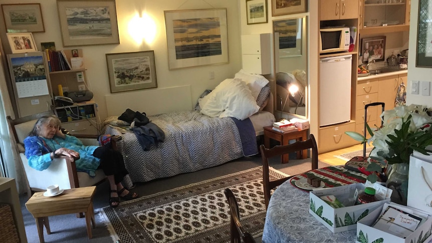 An elderly woman sits in a chair next to a bed in a nursing home room.