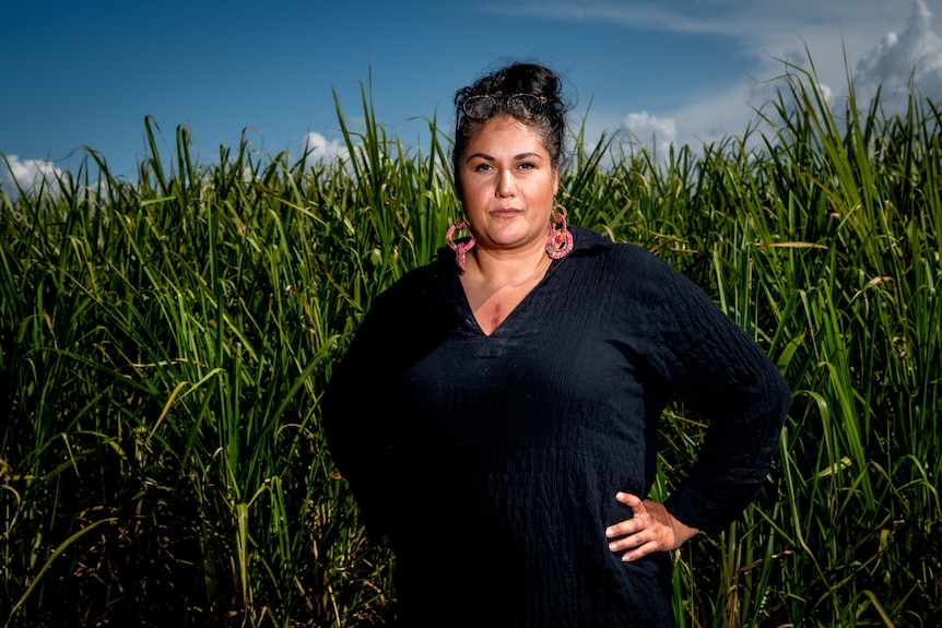 An Aboriginal woman wearing a dark long sleeved shirt and pink earrings standing with her hands on her hips in front of a field