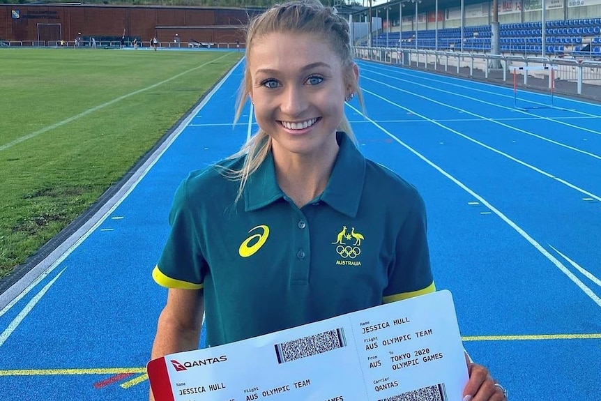 Young woman standing on an athletic track smiling holding a large boarding pass that says she has qualified for Tokyo