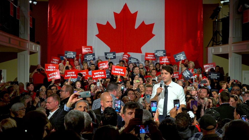 Justin Trudeau talks into a microphone surrounded by a crowd of people in front of a large Canadian flag.