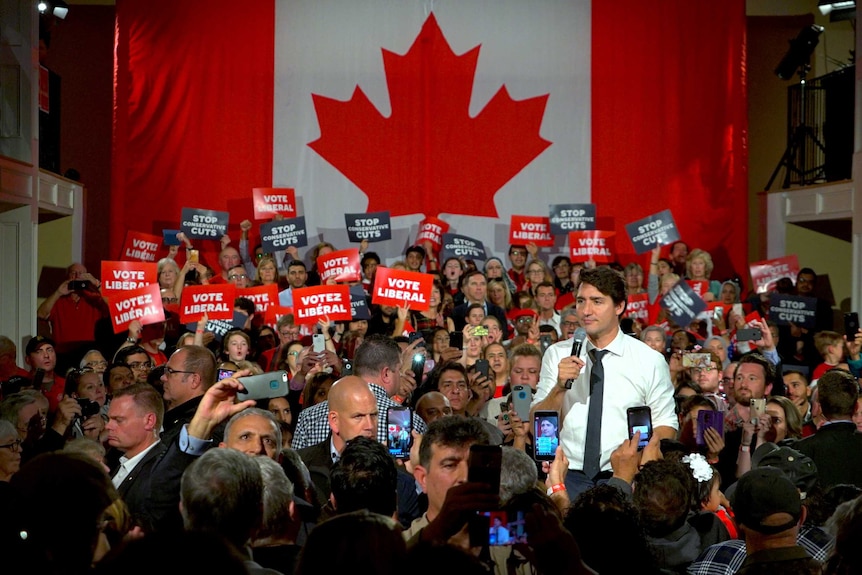 Justin Trudeau talks into a microphone surrounded by a crowd of people in front of a large Canadian flag.