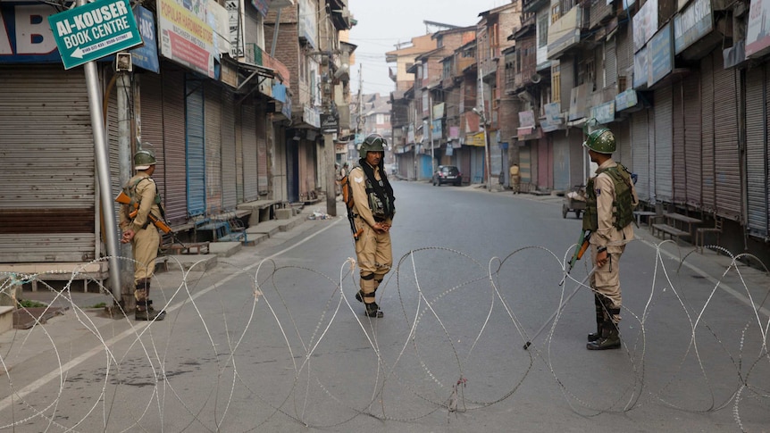 Three armed soldiers stand in the middle of a street of apartments, behind a large barricade of barbed wire.