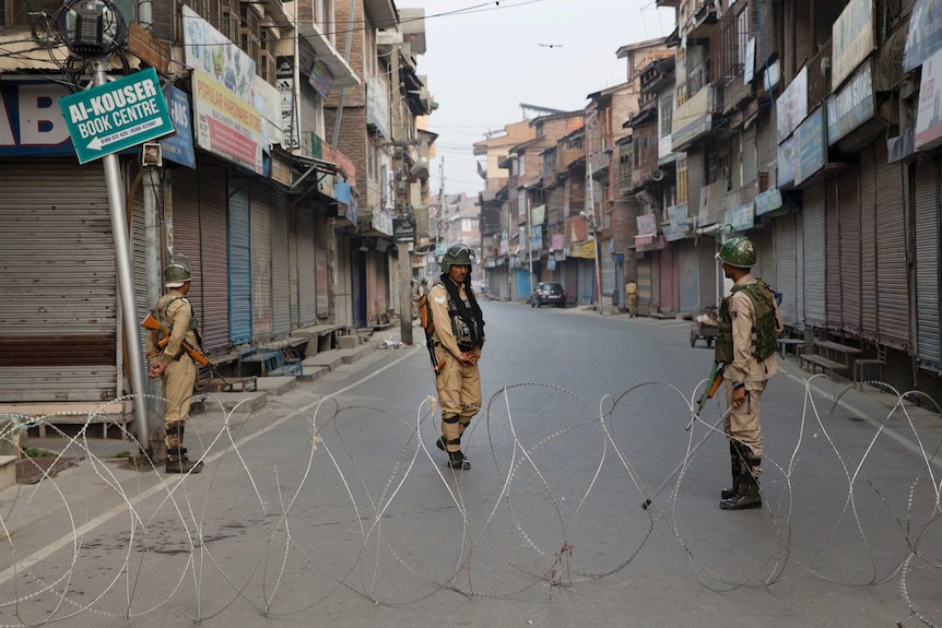 Three armed soldiers stand in the middle of a street of apartments, behind a large barricade of barbed wire.