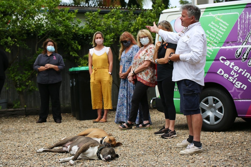 Five women and a man stand outside with two dogs on the ground.