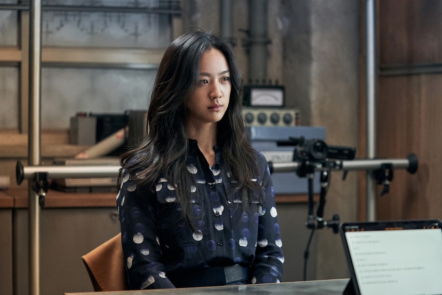 Mid-shot of East Asian woman in dark blue top with white patterns, seated in office looking teary-eyed as if being interrogated