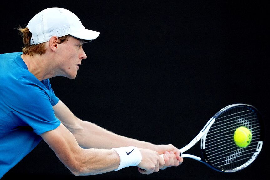 Wysoki blond tenisista w białej czapce i niebieskiej koszuli uderza bekhendem w nocnym meczu tenisa