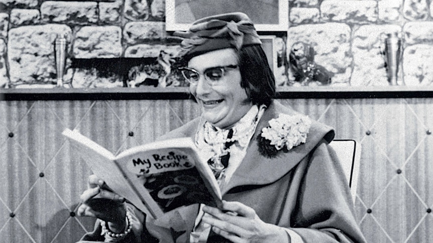 Barry Humphries, appearing as Dame Edna, reads a book on a black and white set