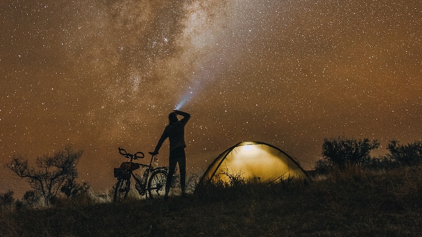 A man holding a bike standing next to an illuminated tent. Above him, the milkyway stretches exapnsively.