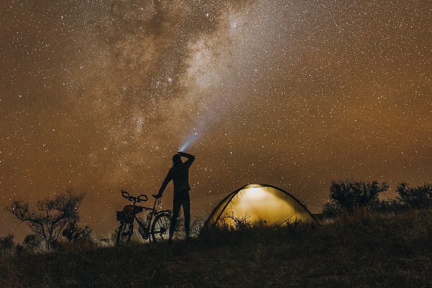 A man holding a bike standing next to an illuminated tent. Above him, the milkyway stretches exapnsively.