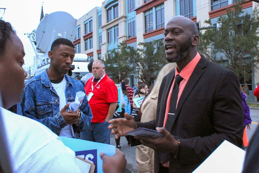 A man in a black suit and pink shirt talking outside a building