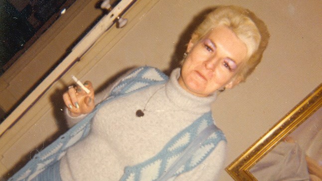 An angled shot of Shirley Finn standing indoors holding a cigarette and wearing a blue outfit and blue bag.