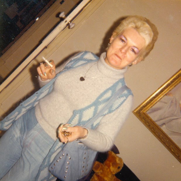 An angled shot of Shirley Finn standing indoors holding a cigarette and wearing a blue outfit and blue bag.