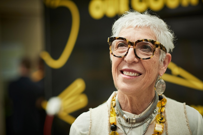 A woman with short gray hair and thick-rimmed glasses smiles as she looks up.