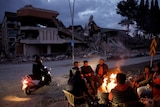 a group of people warm themselves around a fire next to destroyed buildings