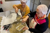 Two lder women wearing aprons and hair nets portion out spaghetti into rectangular foil containers in a commercial kitchen.