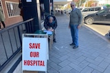 Two elderly men stand on a main street sidewalk. A sign reading "SAVE OUR HOSPITAL" is in the foreground