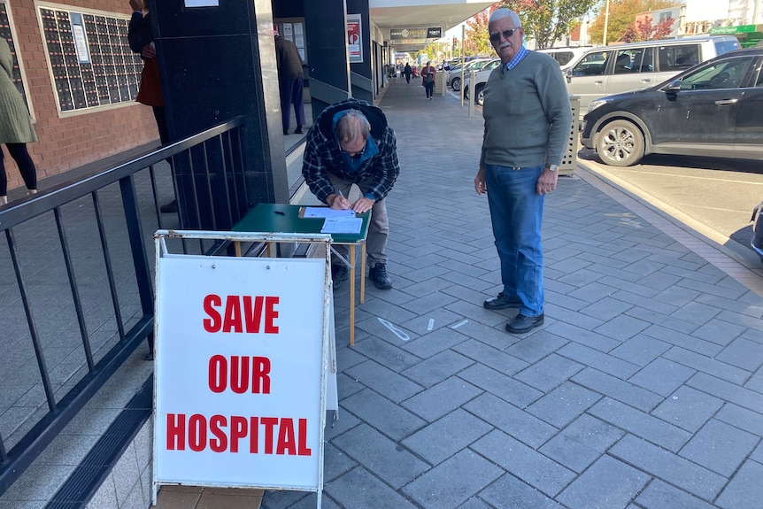 Two elderly men stand on a main street sidewalk. A sign reading "SAVE OUR HOSPITAL" is in the foreground
