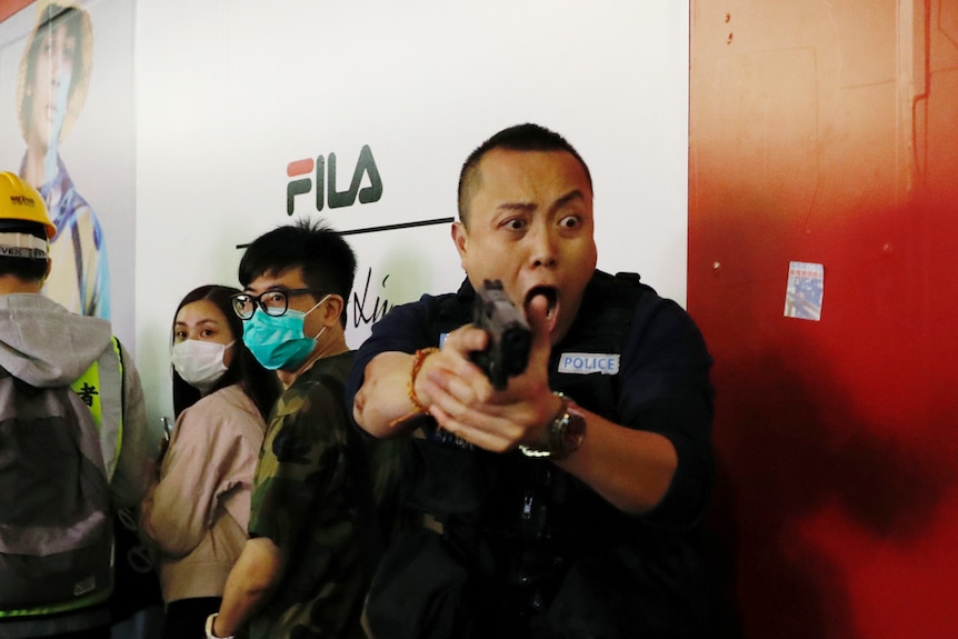 Up against fashion billboard, you see a police officer shouting and pointing his pistol at protesters in face masks.