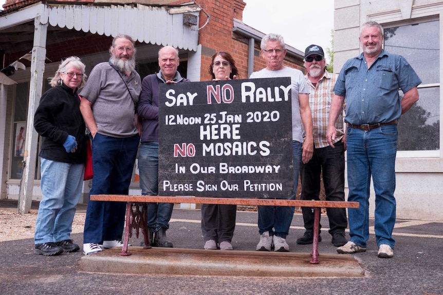 Seven people stand behind a bench with a hand-painted sign that says: say no rally - no mosaics in our broadway'