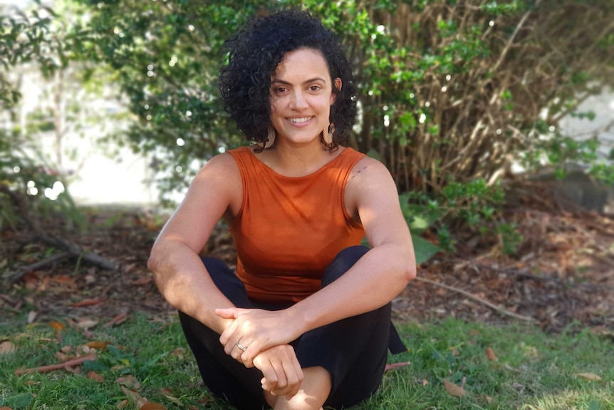 Gabriela Cabral da Rocha Weiss sitting crossed legged, smiling, with shrubs in background.