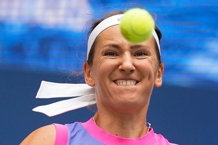 A tennis player grimaces as she swings (racquet unseen) at a ball in front of her face.
