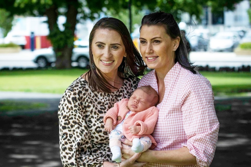 Two women with dark hair hold a baby between them in a park