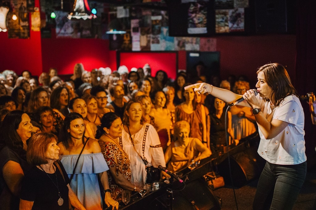 Women stand in a bar singing as a lady stands on stage with a microphone teaching them the arrangement.