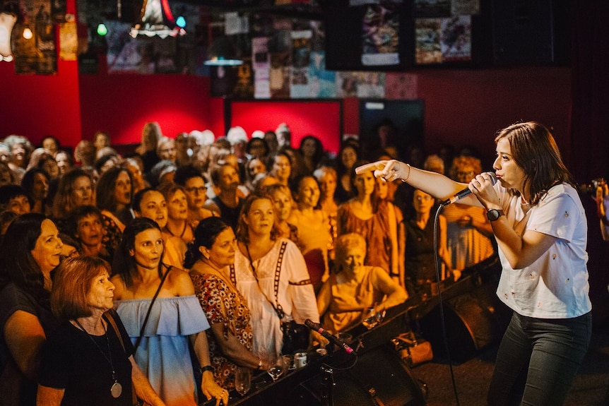 Women stand in a bar singing as a lady stands on stage with a microphone teaching them the arrangement.