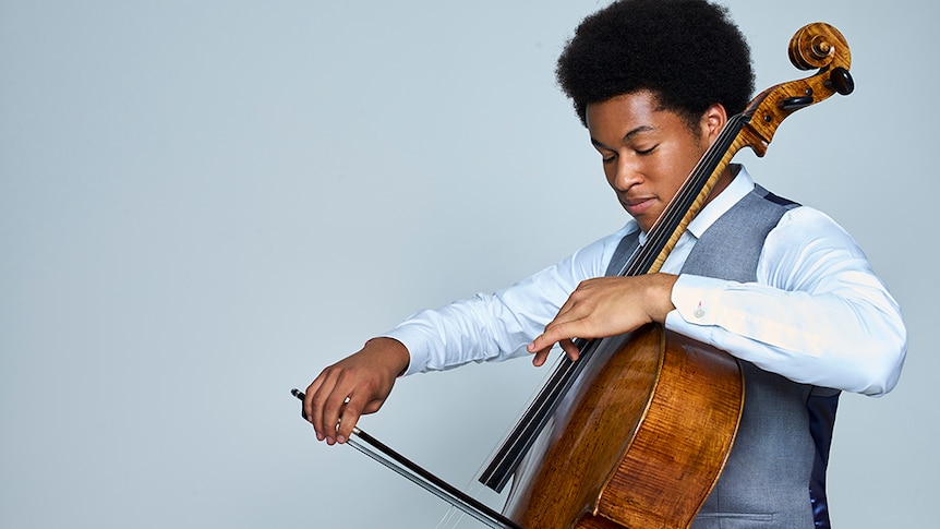 British cellist Sheku Kanneh-Mason plays cello side-on to the camera. He wears a grey vest & white shirt & has his eyes closed.