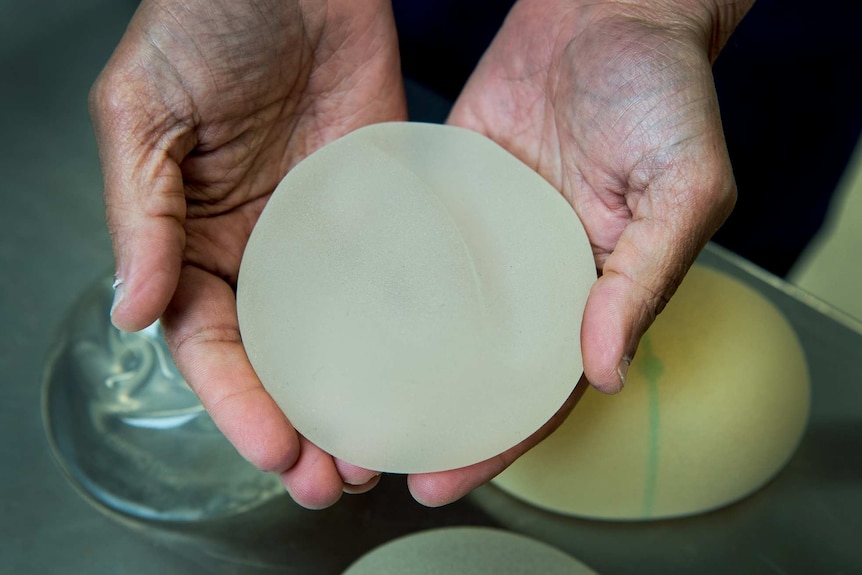 Two hands hold a near-white breast implant above a table.