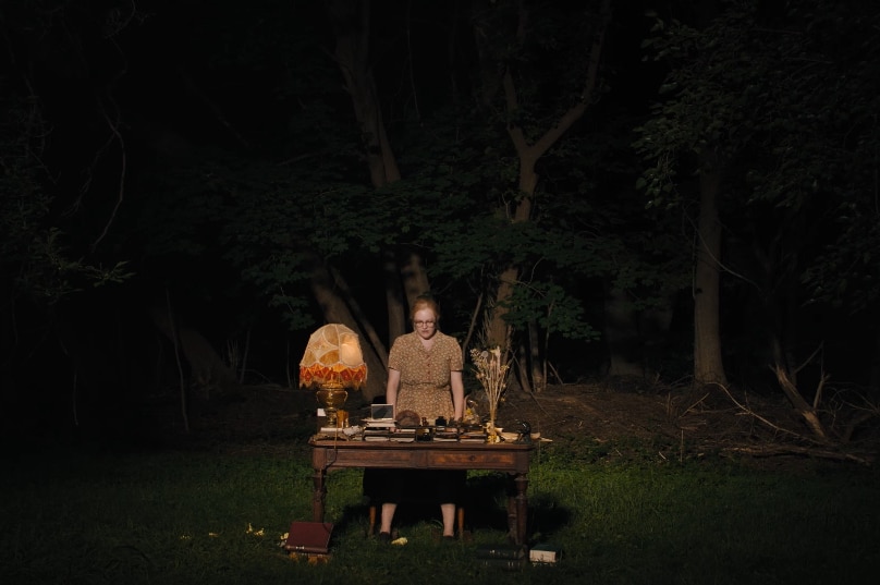 A bespectacled woman in pattern dress stands over wooden desk with lamp, piles of books, papers in forest at night time.